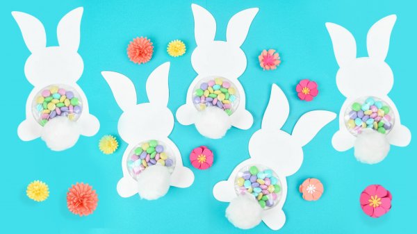 Five white paper 2D bunnies lay on a light-blue background. Each bunny has a plastic dome of candy and a fluffy white cotton tail. You can make these Easter Bunny candy holders too! 