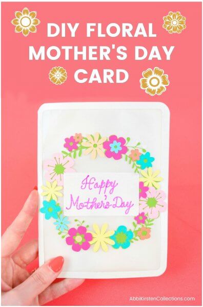 A hand holding a Mother's Day card with a floral wreath on the cover.