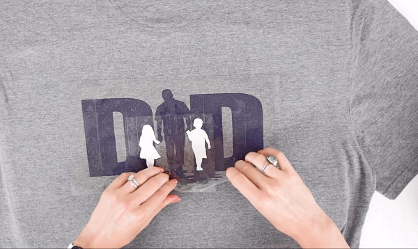 Abbi Kirsten demonstrates how to add a white design layer on top of a darker layer on a gray Father's Day t-shirt. Heat transfer vinyl is used to make the word "Dad" and the silhouettes of children and a father. 