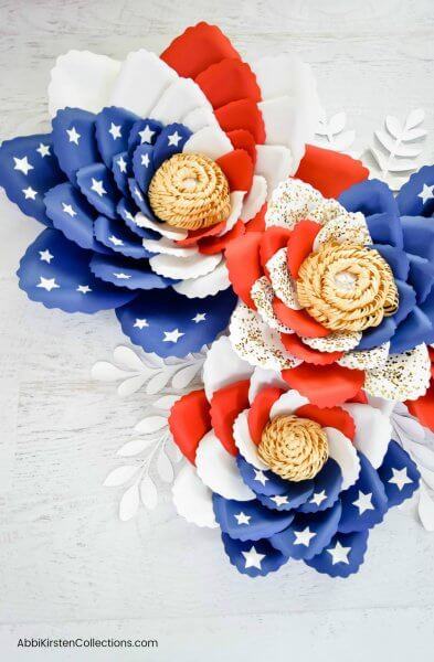 Three giant swirl American flag paper flowers with tan pompom centers and white leaves at the base sit beautifully on a gray wooden table. 