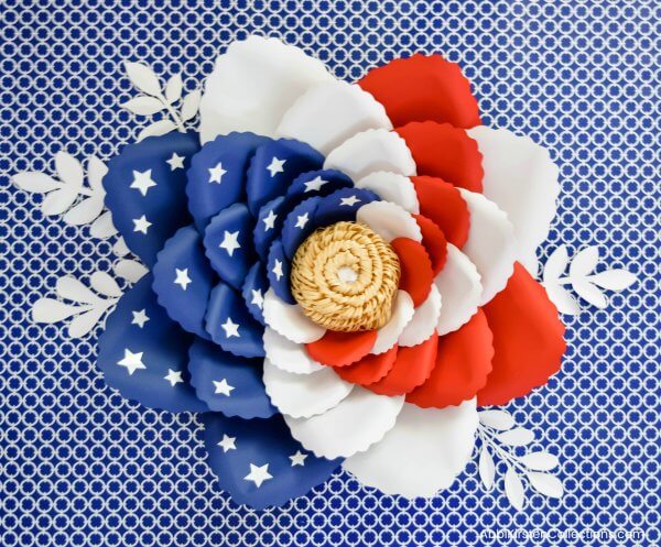 A finished giant swirled American flag paper flower. The red, white, and blue flower petals are swirled around a center pompom, with small white leaves for added foliage. 