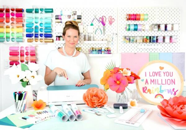 Abbi Kristen is in a white dress standing behind her craft table that holds a Cricut machine, paper flowers, rolls of vinyl, and markers. Behind her colorful craft supplies are organized neatly on shelfs against a large white peg board. 