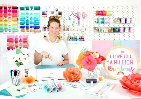 Abbi Kirsten stands with her Cricut machine in her colorful craft room. Her space is organized and full of supplies and giant orange paper flowers. 