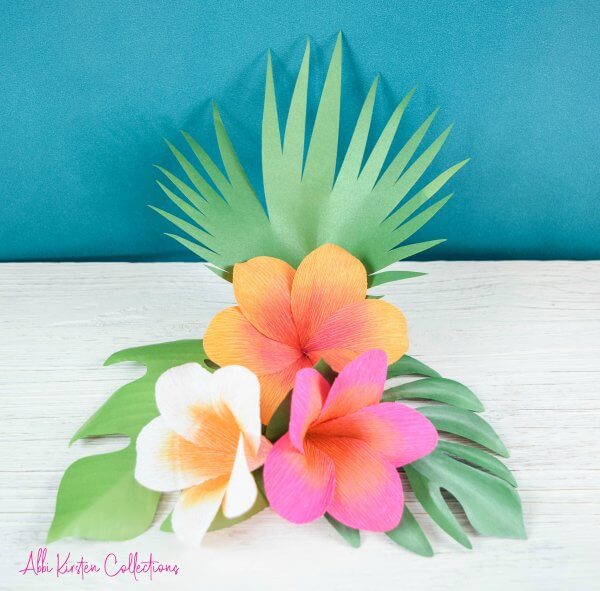 A colorful bunch of orange, white and pink plumeria flowers rest on green leaves on a light grey tabletop with a blue backdrop. "Abbi Kirsten Collections" is written in pink text in the lower left-hand corner. The flowers are made from delicate crepe paper and a Cricut machine. 