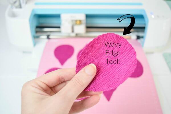 Abbi's hand holds a pink crepe paper petals in front of a Cricut cutting machine with text that says "Wavy edge tool!" 