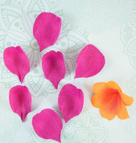 Seven crepe paper pink petals lay next to an orange crepe paper flower on a light green tablecloth with a mandala pattern. How to cut crepe paper with your Cricut Maker. 