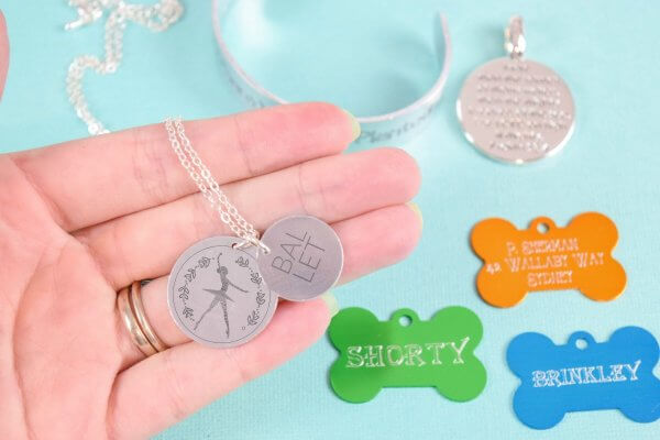 Abbi Kirsten holds a pendant engraved with a ballet dancer. Bracelets and dogs tags, engraved with a Cricut Maker, sit in the background. 