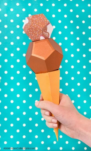 Abbi Kirsten's hand hold a paper ice cream cone with metallic brown ice cream "scoops" in front of a teal backdrop with white polka dots. The secret to this Cricut paper craft is that the 3D ice cream cone holds candy!