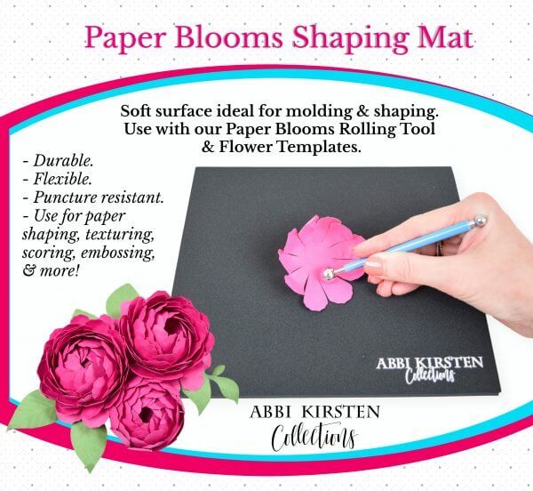 An ad for Paper Blooms shaping mat and tool set, showing a woman's hand using a tool to shape a pink paper flower petal on a black mat, from Abbi Kirsten Collections.