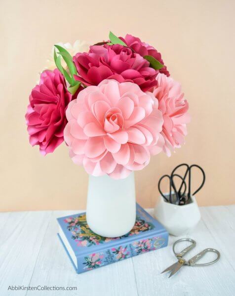A white vase of paper Camellia roses in varying shades of pink with green paper leaves. The white vase is on a blue book, against a beige wall, and next to a white cup full of scissors. 