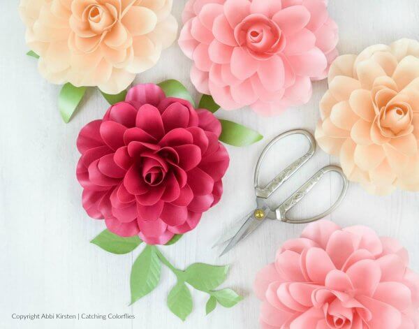 An overhead view of five delicate paper Camellia roses in shades of pink and peach with delicate green leaves, laid out on a white tabletop next to crafting scissors. 