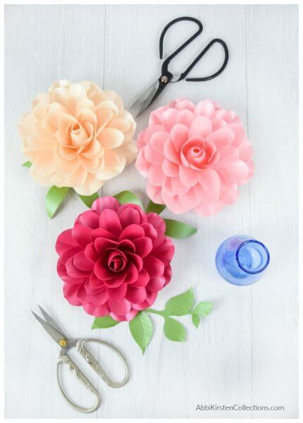 Three paper Camellia roses in shades of pink and peach are seen from above, laying on a white surface along with two pairs of crafting scissors and a small blue glass flower vase.
