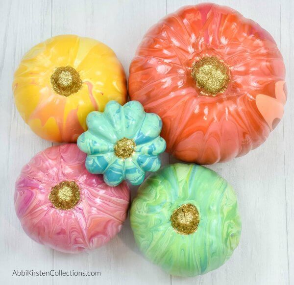Varying sizes of  yellow, orange, green, pink and blue marble painted pumpkins on a white wooden table. 
