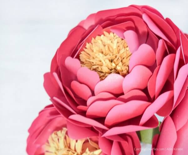 A close-up of pink ombre paper peonies with a yellow paper center and a green stem on a white background. You can learn how to make these easy paper flowers. 
