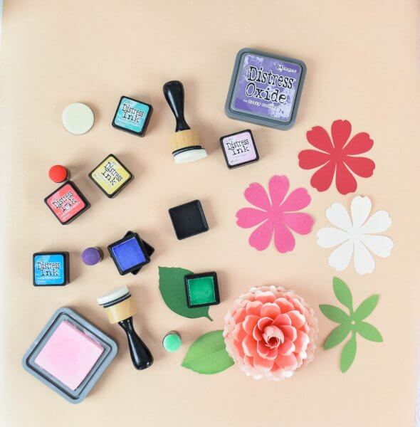 An overhead view of paper flower distressing tools including ink trays, rounded sponges with wooden handles,  pink, red and white petals and green leaves. 