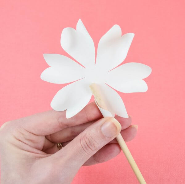 Abbi's hand demonstrates how to curl paper flower petals, using a wood dowel and white paper petals in a salmon background. 