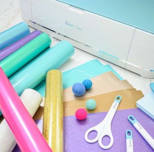 A close-up of a Cricut maker machine, with rolls of vinyl in pastel colors, pom-poms, and various Cricut tools on top of bright cardstock. 