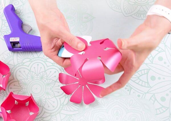 Cricut scraper is used by Abbi Kirsten to curl metallic pink paper flower petals. Finished petals lay on the decorative table next to a purple glue gun. 