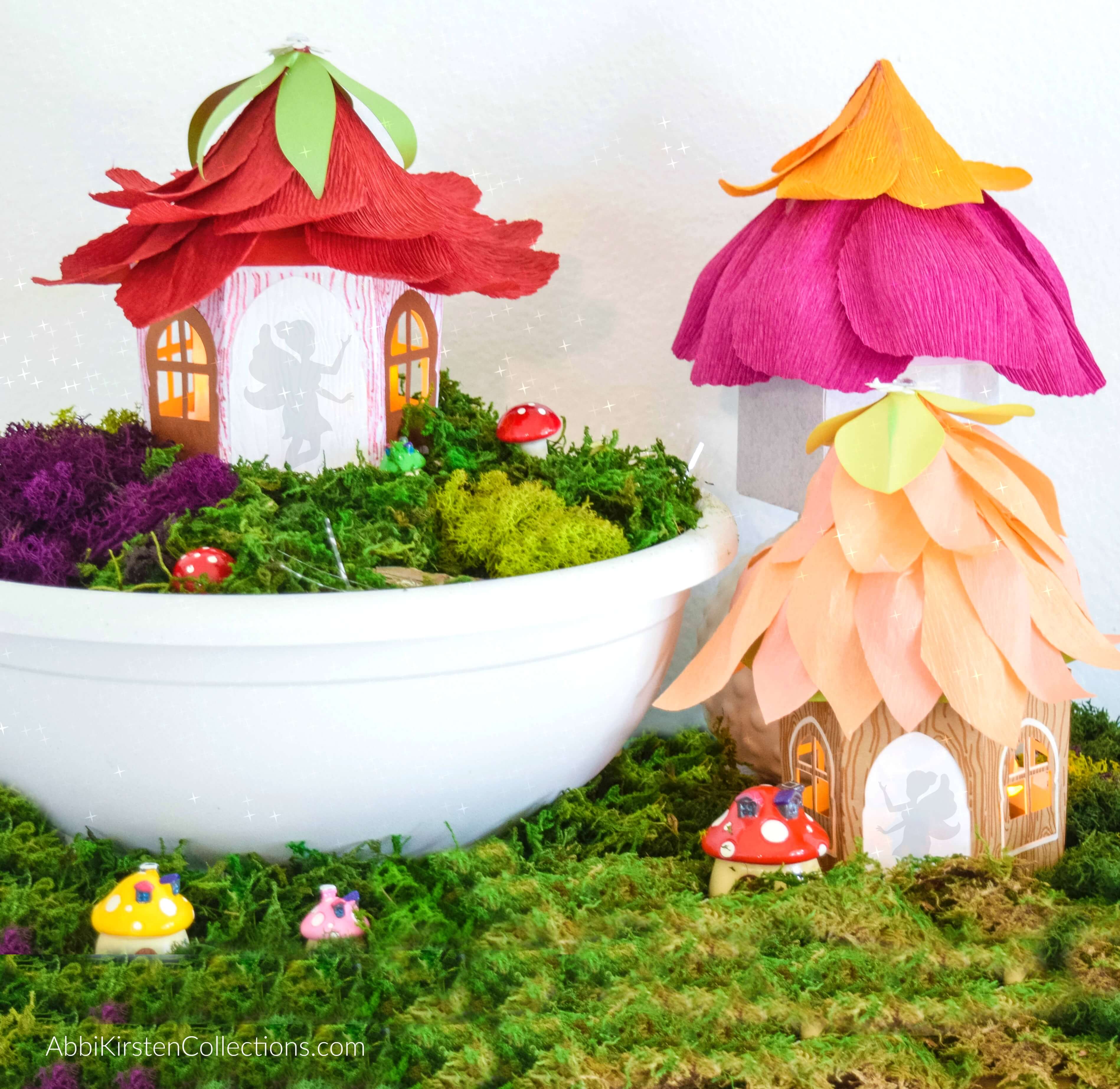 Three paper fairy homes sit in faux moss and a white bowl. The roofs are made of crepe paper to resemble upside-down flowers. The silhouette of a fairy is seen in each door, and small mushrooms adorn the greenery. You can learn how to make these cute houses at AbbiKirstenCollections.com. 