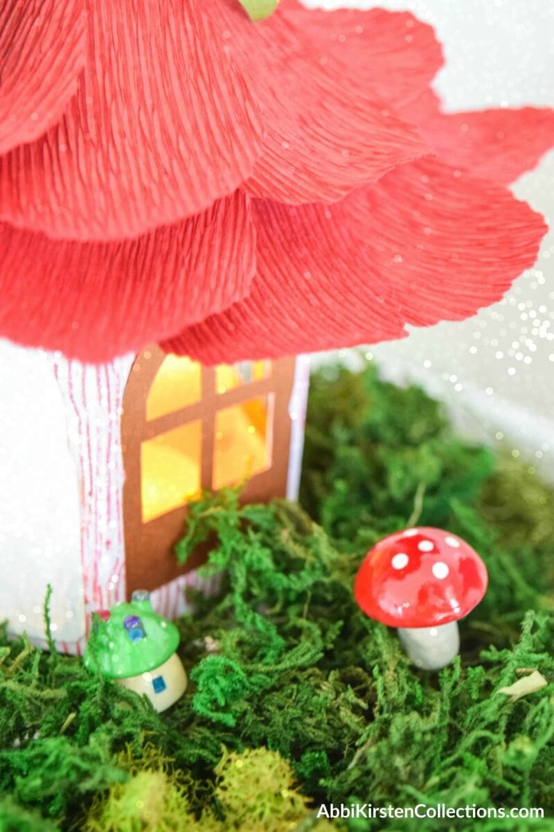 A closeup image of the window in a paper fairy house with crepe paper flower petals for a roof. A warm yellow light spills onto the moss and small mushrooms garden in front of the house. 