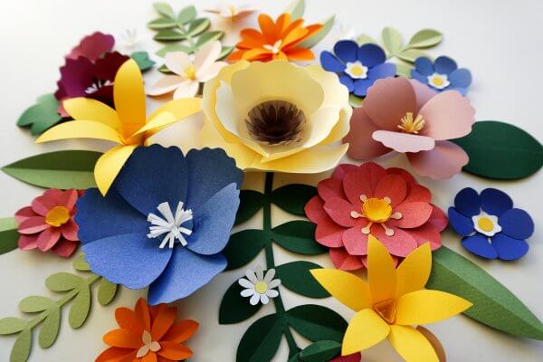 A variety of paper flowers like lilies, daisies, and more in a rainbow of colors surrounded by green leaves are arranged on a white table. 