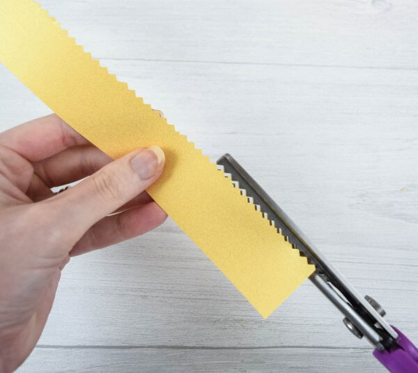 A close-up of Abbi's hands using pinking shears to create a paper flower center on a strip of yellow cardstock. 