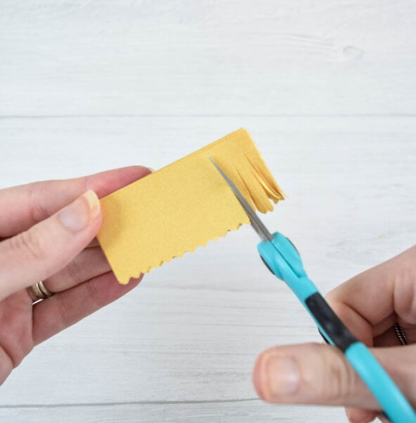 Abbi's hands use detail scissors to cut long strips in a yellow strip of cardstock to be used as the stamen in a paper flower design. 