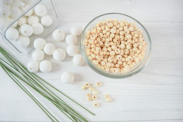 A white wood table holds a glass bowl of wooden balls, as a clear container spills it's supply of white balls next to green wire. These are some supplies you may need to create fantastic paper flowers. 