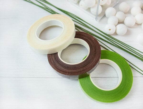 Corsage tape for paper flowers in white, brown and green, with wire stems and white foam balls all on a white table-top. These are some materials you may need for creating paper flowers. 