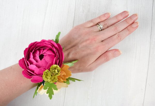 A picture of a woman's arm over a white wooden table, with a pink and yellow paper flower corsage om a slap band. 