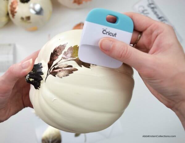 Abbi Kirsten holds a small white pumpkin in her hand as she uses a scraper to burnish textured, metallic vinyl leaves onto the faux pumpkin. These elegant pumpkins will class up any Halloween or Autumn décor. 