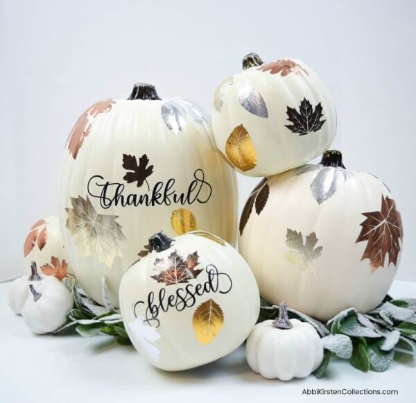 White pumpkins are stacked together with words and textured metallic leaves.  Snowy leaves decorate the floor. 