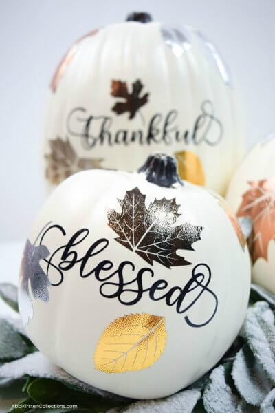 Two faux white pumpkins, one in the background and one in the foreground, have been decorated with metallic vinyl leaves and Autumn sayings like "Blessed" and "Thankful."
