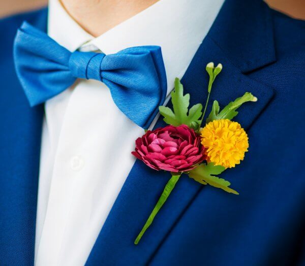 A close-up of a red and yellow paper boutonniere with green leaves pinned on a deep blue suit with a blue bowtie on a white shirt.