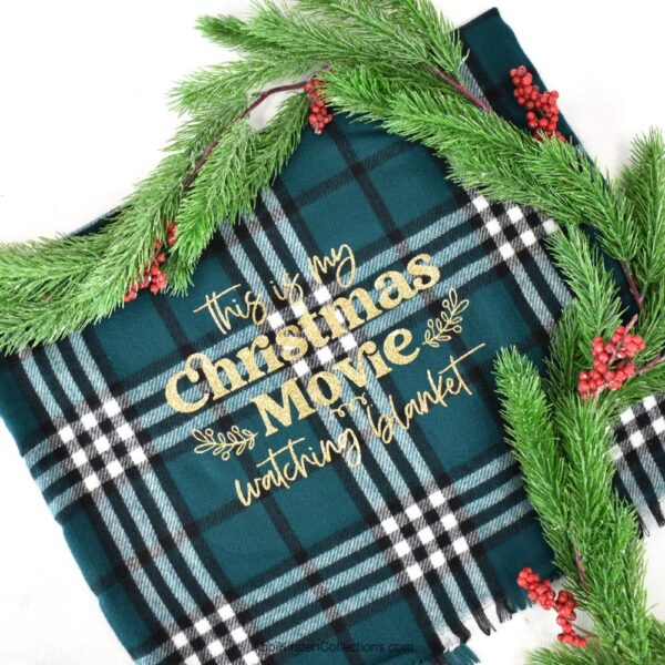 The images shows a Christmas blanket in plaid green with the phrase "this is my Christmas movie watching blanket" in gold glitter heat transfer vinyl. Download the free Christmas blanket SVG file. 