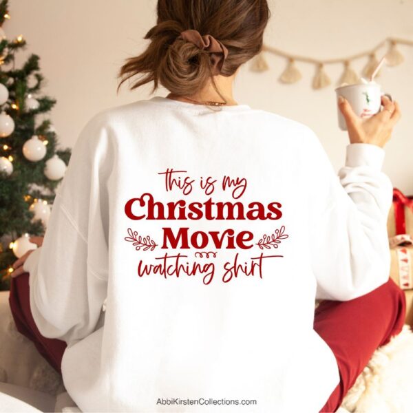 The image shows a girl wearing a white sweatshirt that says, "This is my Christmas movie watching shirt" in red iron-on vinyl. She's holding a mug of coffee and sitting by a Christmas tree.