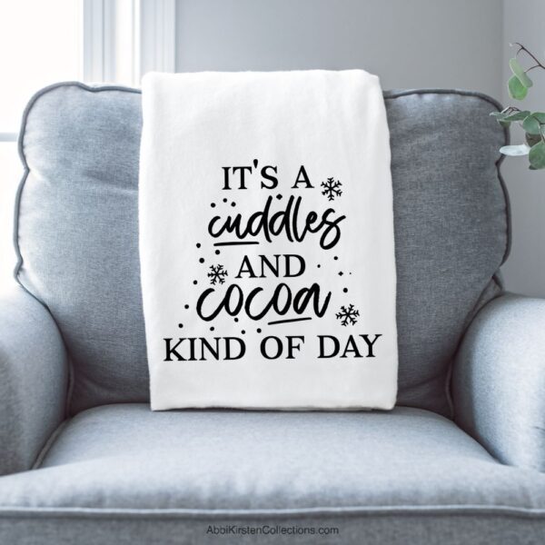 A white throw blanket hanging over the back of a blue arm chair. The blanket has a black iron-on vinyl decal that says "it's a cuddles and cocoa kind of day" surrounded by snowflakes.