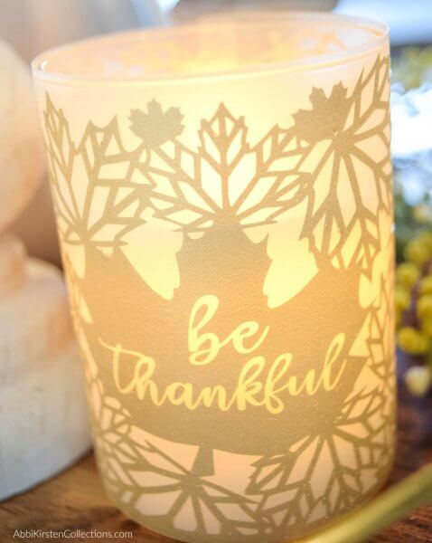 A glass candle jar with a flickering LED candle flame is wrapped in a golden fall leaf design candle wrap. A large leaf in the center of the jar has the words “be thankful” carved into the vinyl.