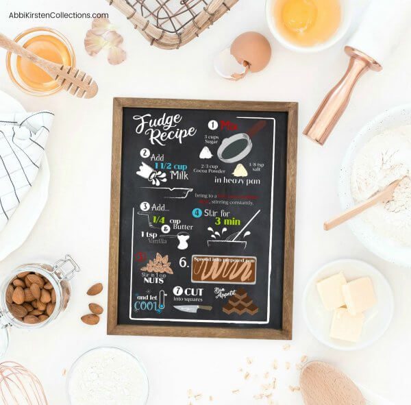 An overhead image of a black sign with a fudge recipe in white text and colorful baking images. The sign is surrounded by baking ingredients and tools on a white workstation. 