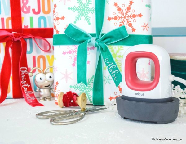 Christmas presents wrapped in green and red ribbons sit next to scissors, ribbons and a Cricut EasyPress and a toy bug. Use the Cricut Easypress Mini and iron-on vinyl to customize gift bows with your recipient's name. 