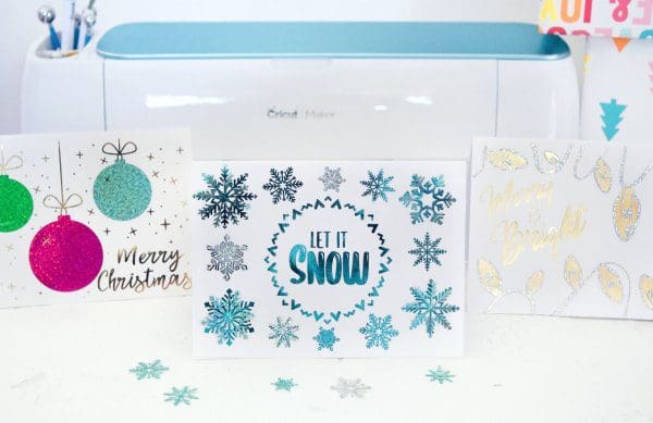 Three Christmas cards made with free templates stand in front of a Cricut cutting machine. Blue snowflake confetti is sprinkled around. The card on the left says "Merry Christmas" with purple, blue and green ornaments. The front card says "Let it Snow" in blue letters surrounded by blue snowflakes. 