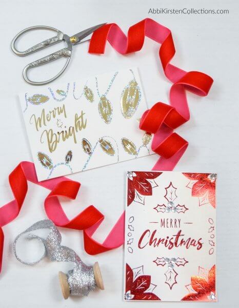 Two Christmas cards, red and silver ribbon, and scissors adorn the gray table top. The cards read "Merry and Bright" and "Merry Christmas"