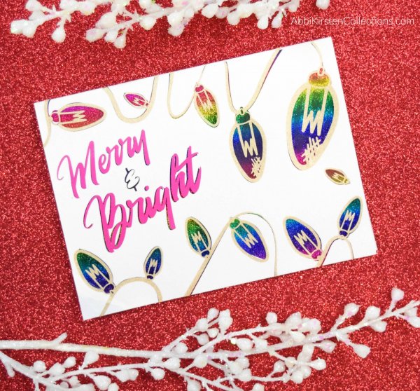 A colorful Christmas card decorated with rainbow deco foil Christmas lights reads "Merry and Bright" in pink cursive letters. The card lies on a red glittery background. 