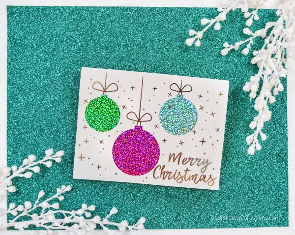 Glittery, colorful ornaments adorn the front of this DIY Christmas card template available for free! The card rests on a green glitter background with white flower buds in the corners. 
