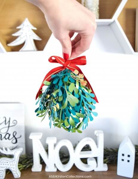 A green paper mistletoe kissing ball topped with a red and gold ribbon hanging above all white Christmas decorations atop a wooden table. 