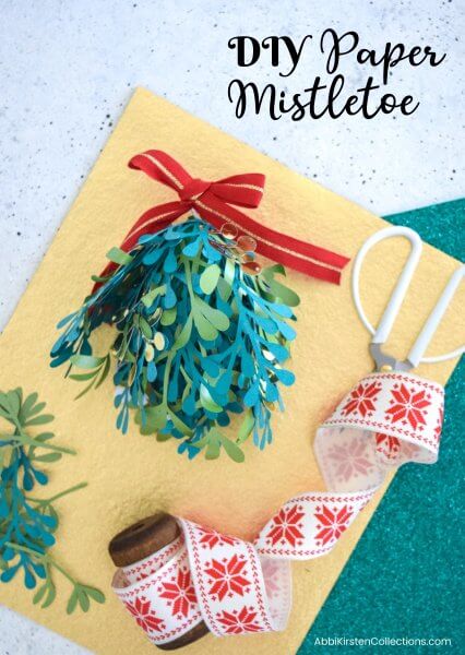 A festive green paper mistletoe Christmas kissing ball sits on top of yellow construction paper, surrounded by Christmas ribbon and crafting supplies. This tutorial and free SVG mistletoe template make the holiday crafting easy and fun!