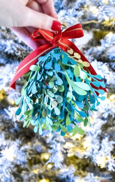 This finished paper mistletoe Christmas kissing ball is elegant and looks incredibly festive, held up in front of a decorated Christmas tree with white and gold lights.