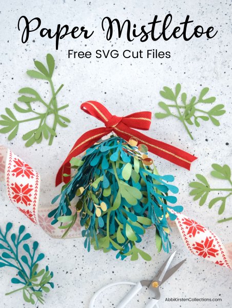 A green paper mistletoe Christmas Kissing Ball alongside red and white snowflake ribbon and small white trimming scissors atop a white and black speckled counter. 