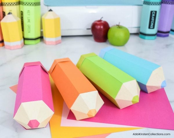 Blank colored pencil foldable boxes are in the foreground, with finished paper crayon and pencil shaped boxes in the background. Crafts are surrounded by red and green apples and colorful cardstock. 
