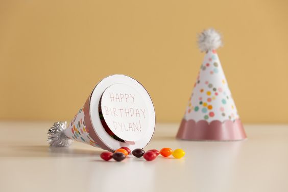 Birthday hat favors against a mustard yellow background. One hat is on it's side revealing the engraved paper bottom reading "Happy Birthday Dylan!"